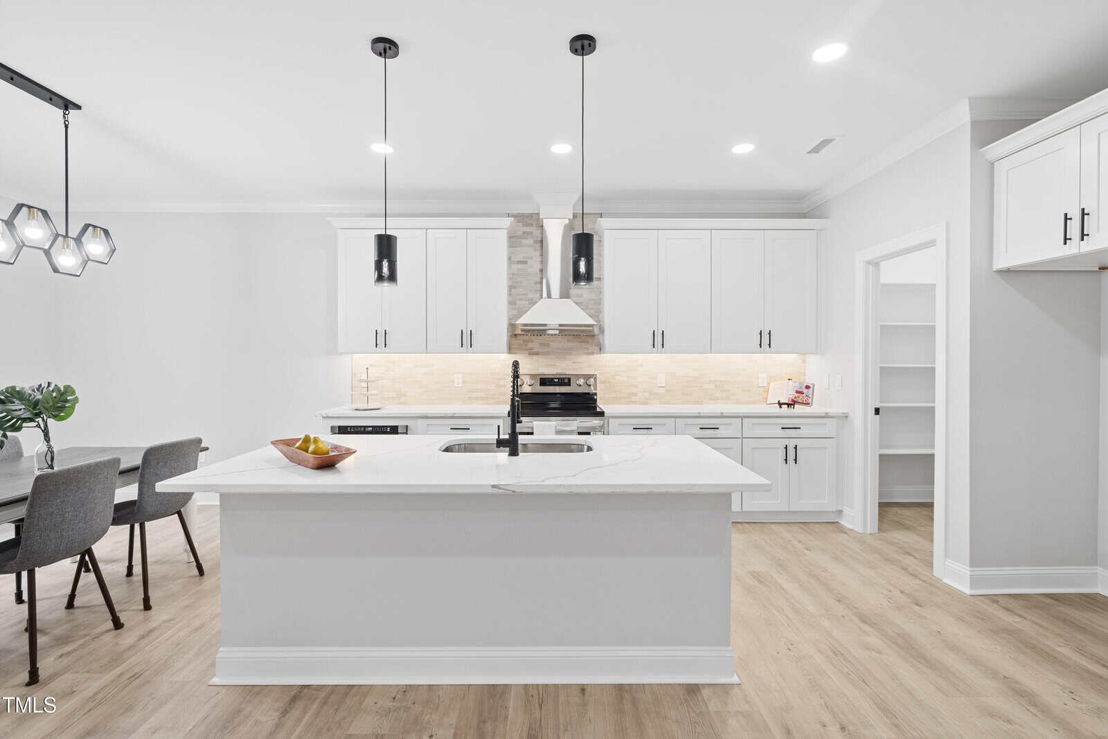 Modern kitchen with white cabinets, a large island with a marble countertop, two black pendant lights, wood flooring, and stainless steel appliances. Dining area with a table and chairs is visible to the left.