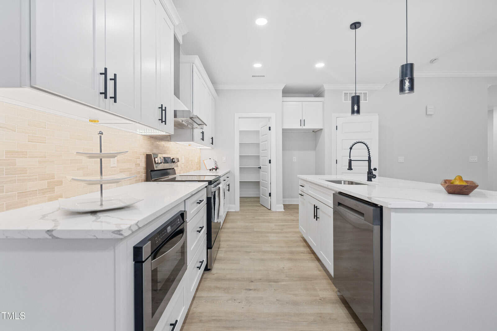 Modern white kitchen with stainless steel appliances, marble countertops, light wood flooring, a center island with a black faucet, and a walk-in pantry with an open door in the background.
