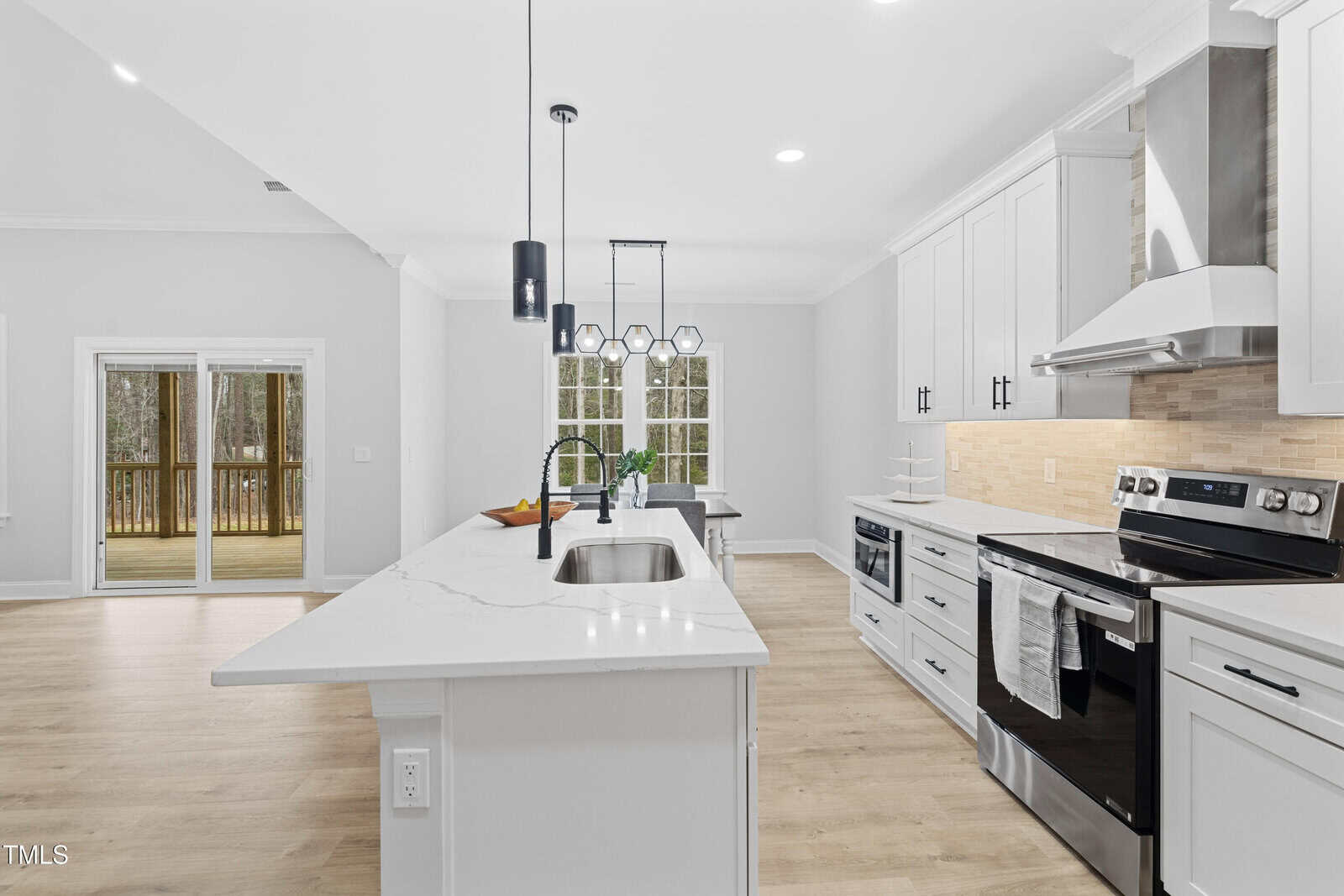 Modern kitchen with white cabinets, stainless steel appliances, marble countertop island with a sink, pendant lights, light wood flooring, sliding glass doors, and a window facing a wooded area.