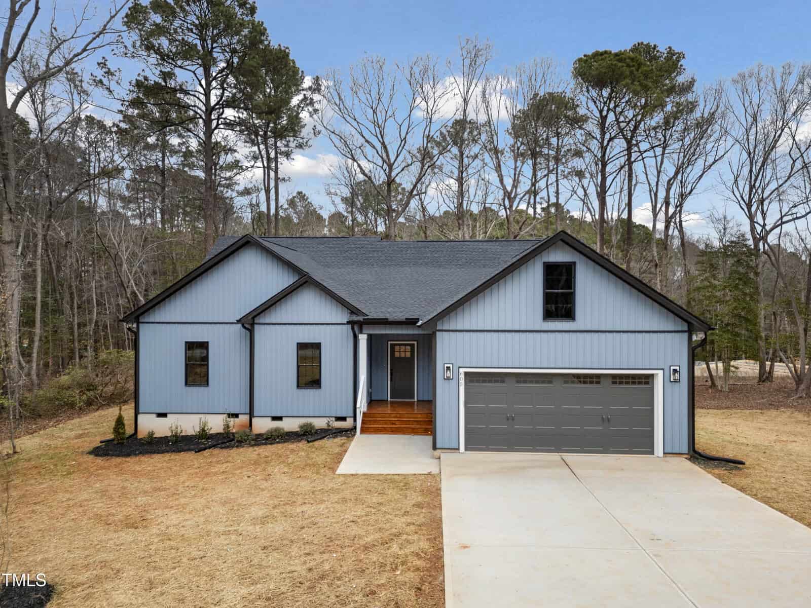 A modern blue single-story house with a black roof, double garage doors, central entrance, and wood steps. The house is surrounded by leafless trees and sits on a large, brown lawn with a concrete driveway.