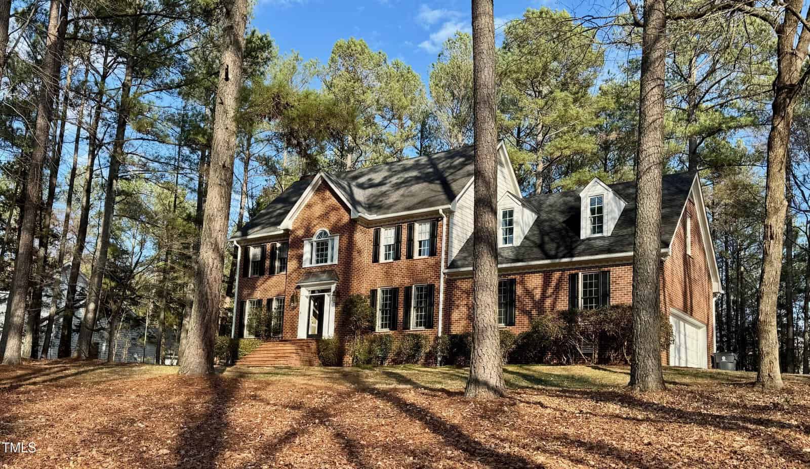 A two-story brick house with a steeply pitched roof is surrounded by tall trees. The home features multiple windows and a white-trimmed front door. Fallen leaves cover the ground, indicating a fall setting.