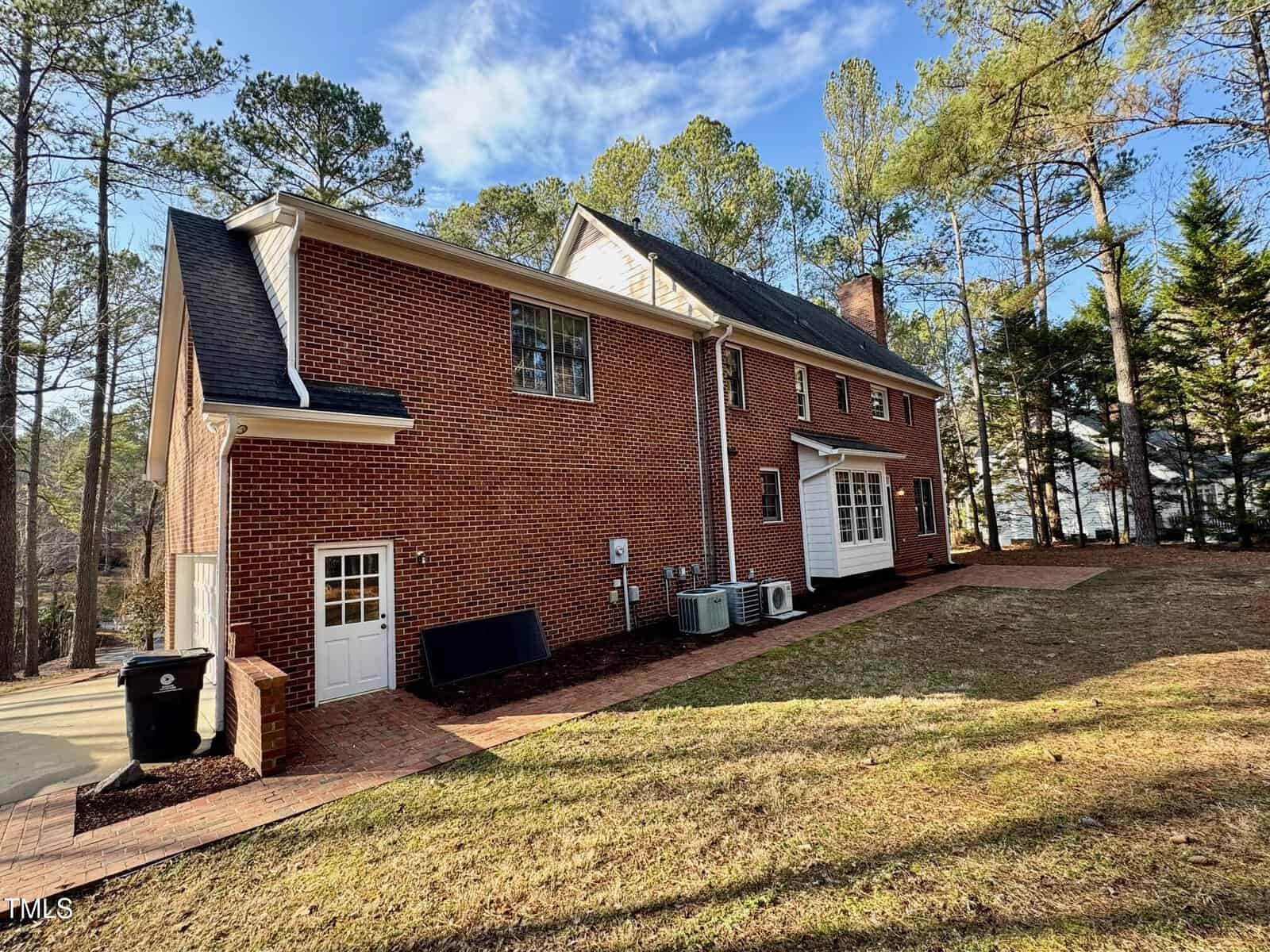 A red-brick two-story house with a black shingle roof, surrounded by trees. The side view shows several windows, a white door, air conditioning units, a trash bin, and a paved walkway leading around the house. The sky is clear with scattered clouds.