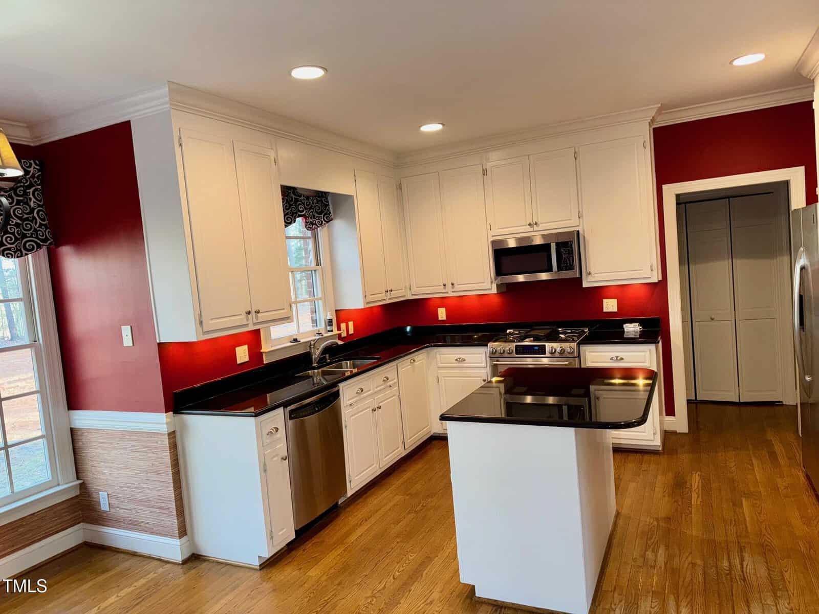 A kitchen with white cabinets and a black countertop. It includes a stove, oven, dishwasher, and microwave. The walls are red, with wooden flooring. Recessed lighting is installed in the ceiling, and there are two windows with patterned valances.