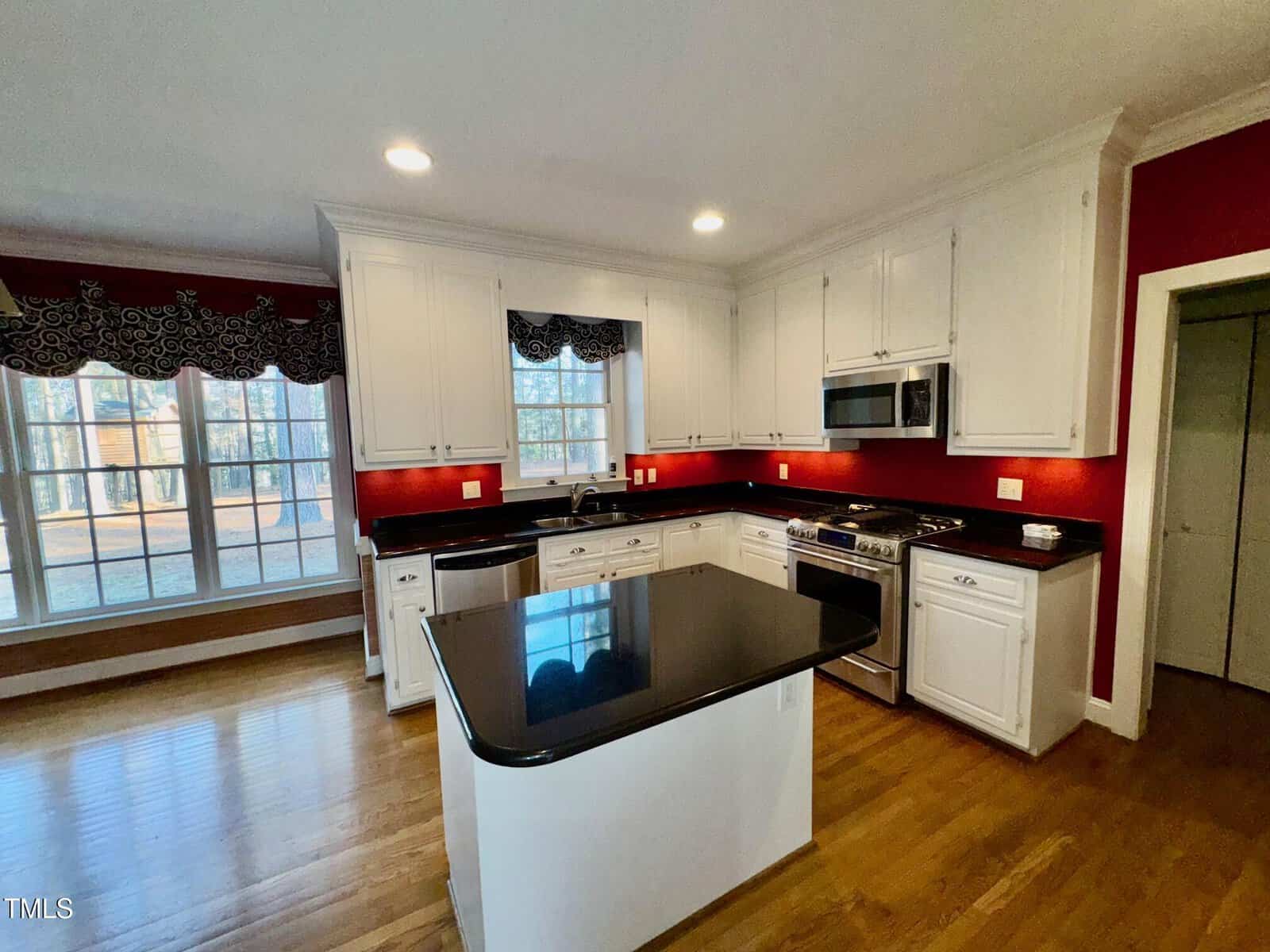 A modern kitchen with white cabinets, stainless steel appliances, and a black granite countertop island. The walls are painted red, and there are large windows with patterned valances. The floor is wooden, and there is recessed lighting.