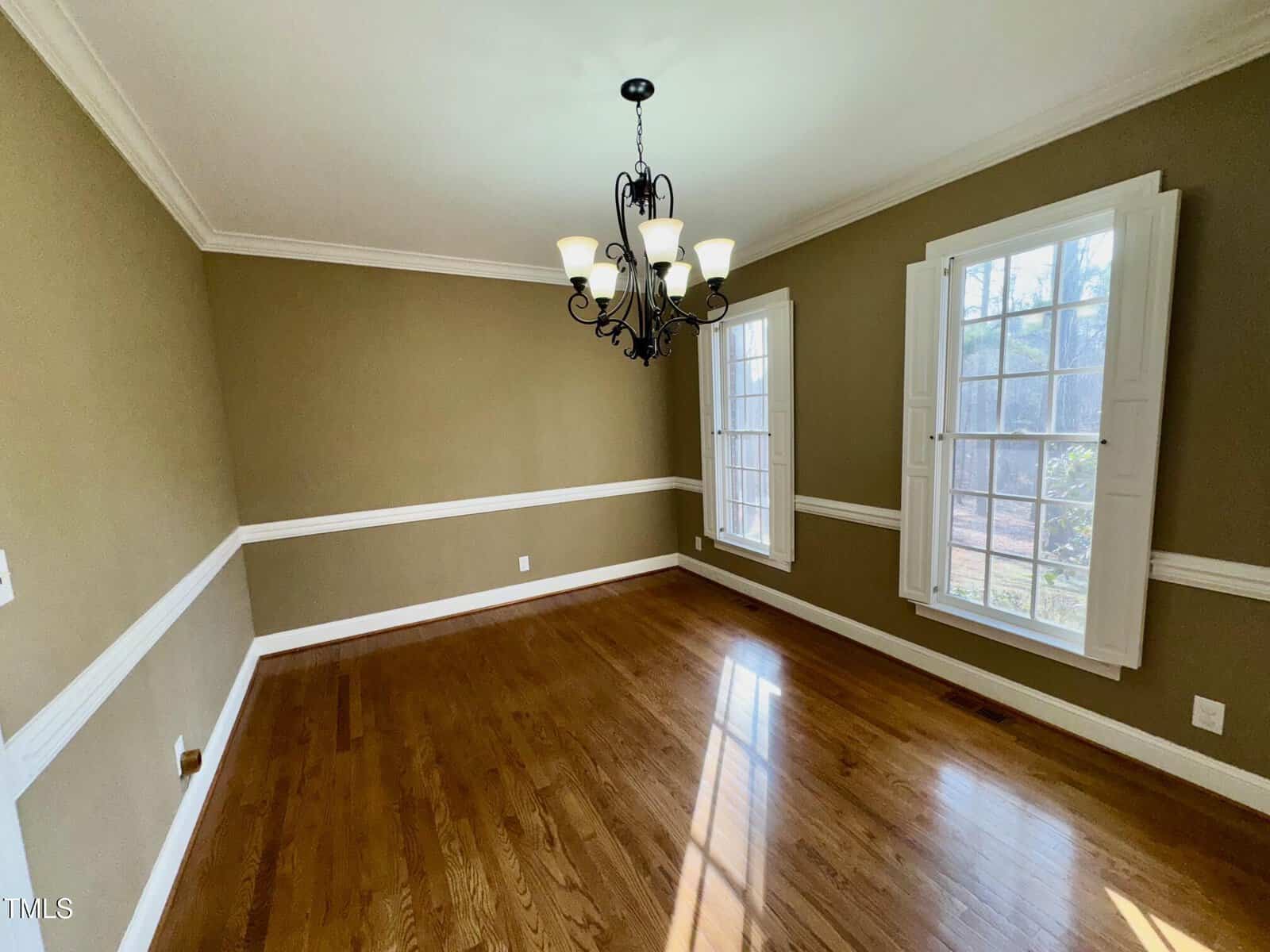 A dining room with hardwood floors, tan walls, and white trim. Two large windows with white shutters let in natural light. A black chandelier with multiple bulbs hangs from the ceiling. The room is empty of furniture.
