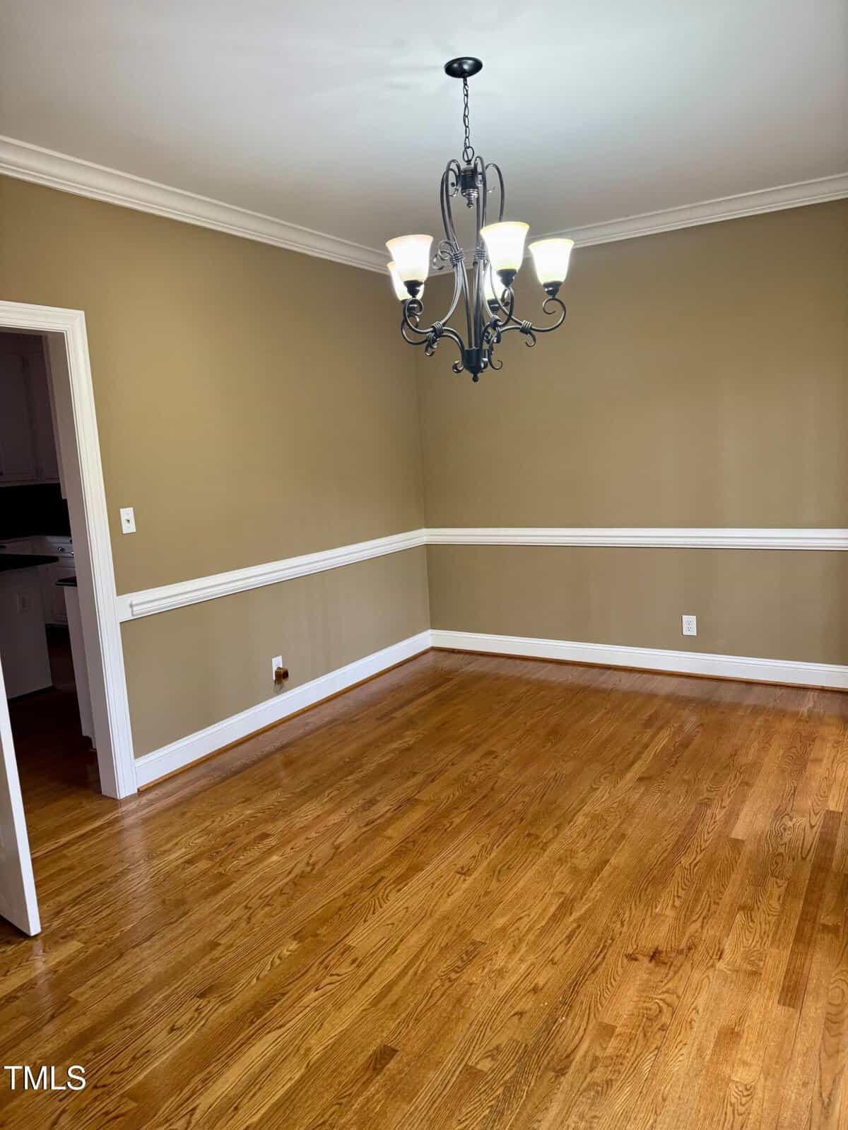 Dining room with tan walls and white trim, featuring a dark metal chandelier with five lights. The floor is hardwood, and there's a white door open on the left side leading to another room.