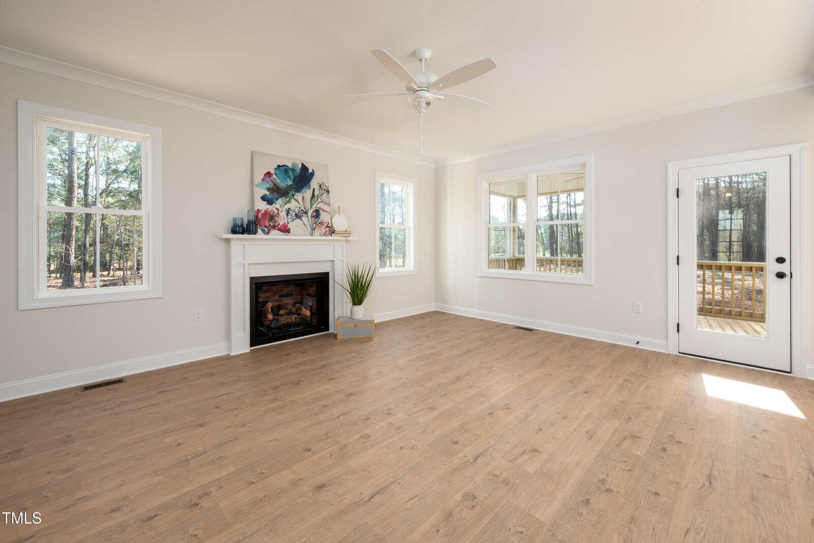 A bright, empty living room with light wood flooring, a ceiling fan, three large windows, a glass door, a white fireplace with art above it, and a potted plant on the hearth. Trees are visible outside.