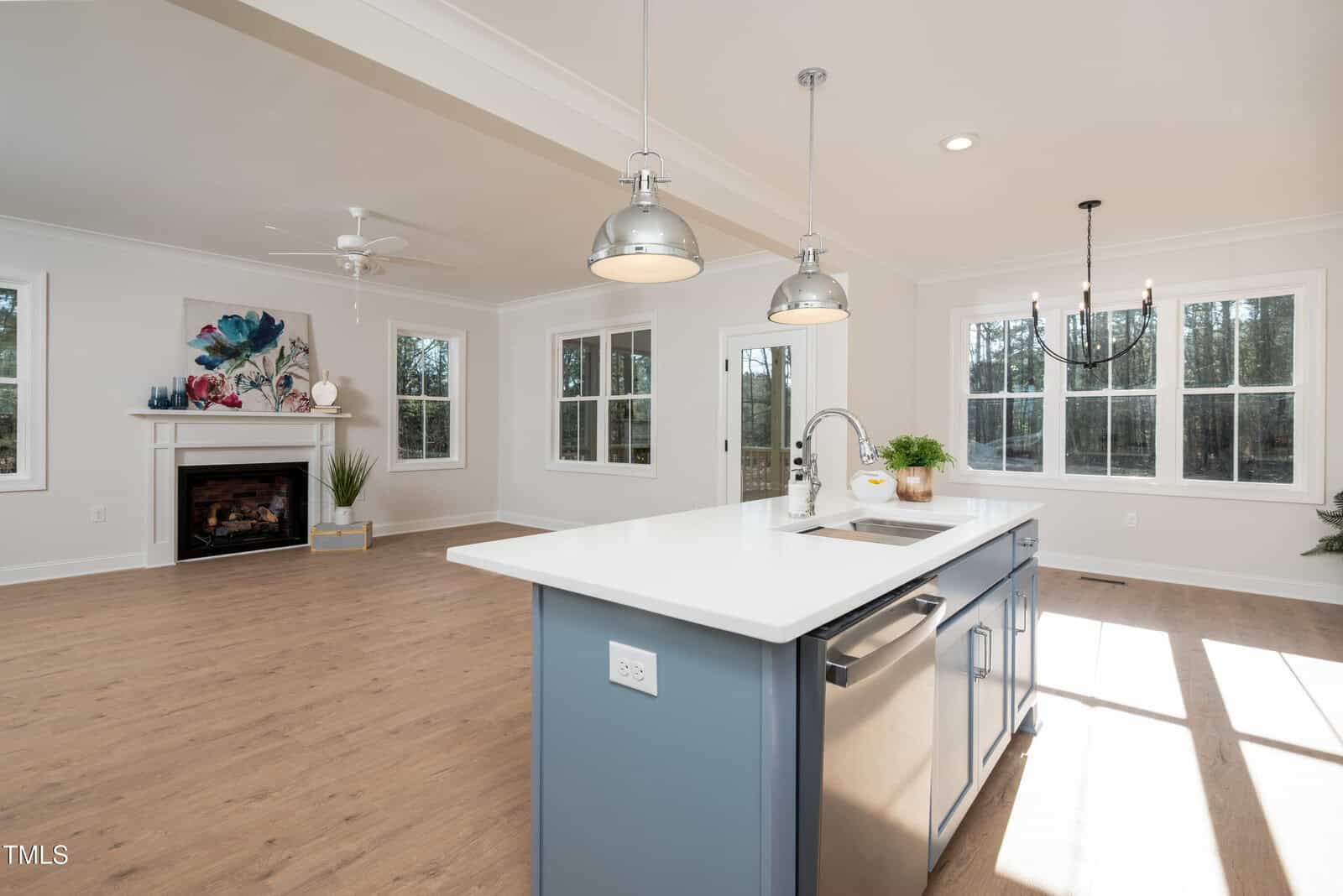 Modern open-concept kitchen and living area with wood floors, blue island, white countertops, stainless appliances, fireplace, ceiling fan, and large windows letting in natural light.
