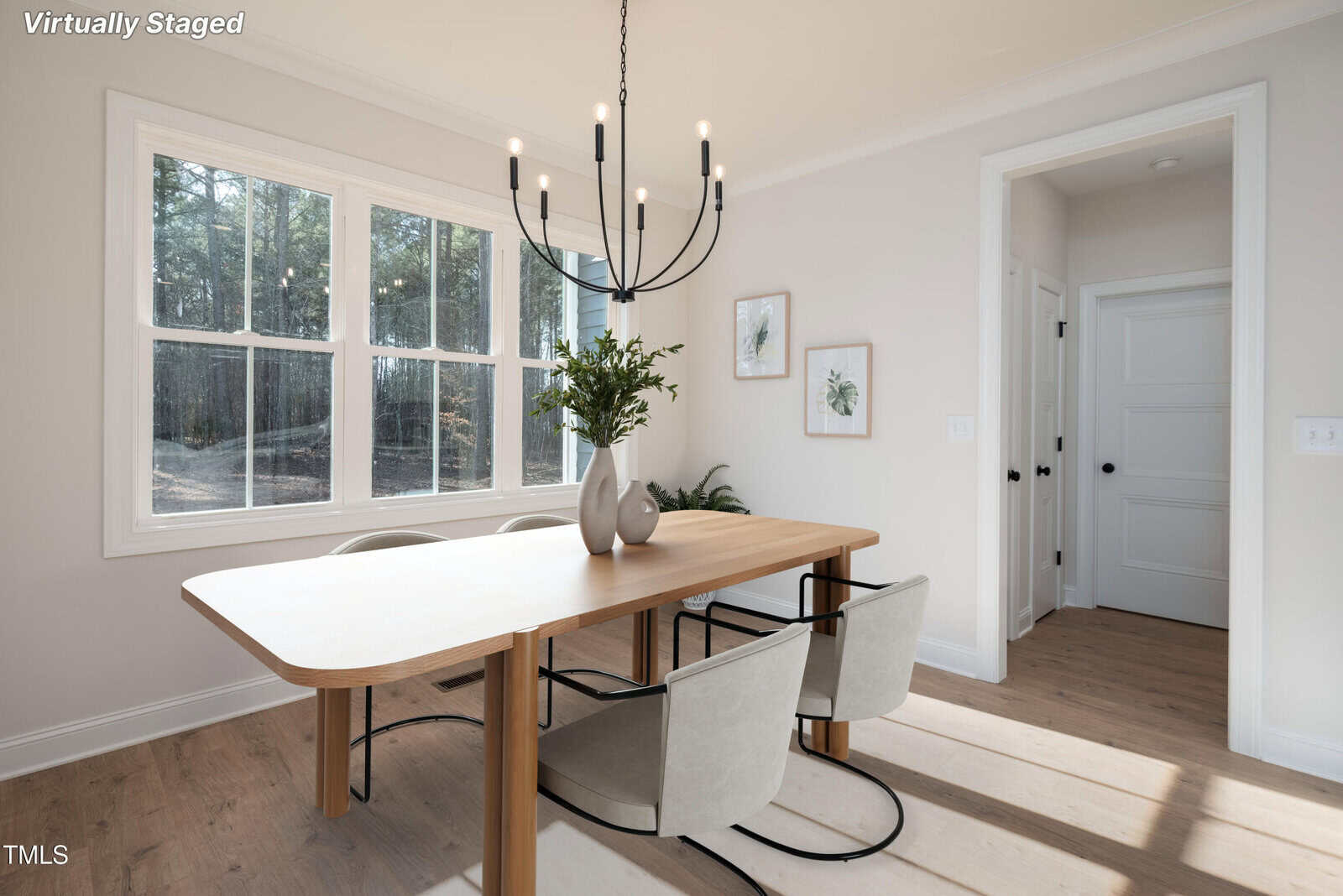 Modern dining area with a light wood table, four beige chairs, a black chandelier, vases with greenery, and wall art. Sunlight streams through large windows, illuminating the bright, minimalist space.