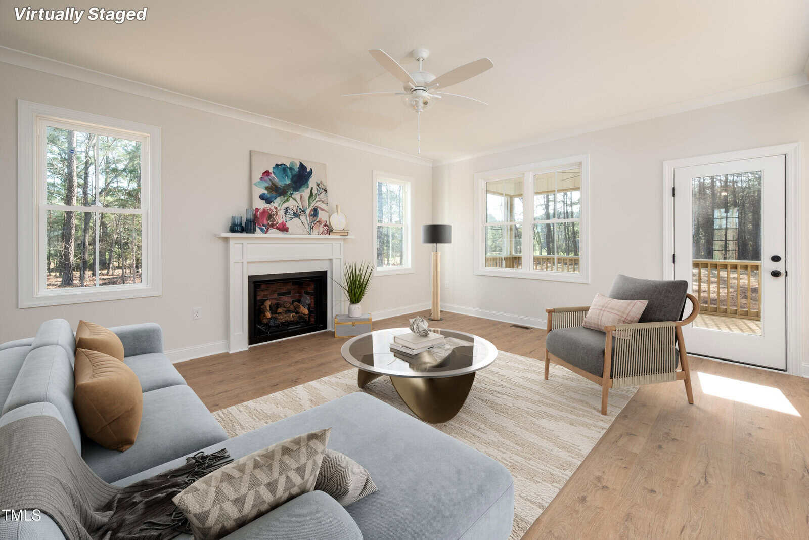 A bright living room with wood flooring, a gray sofa, an armchair, a round coffee table, and a fireplace with art above it. Large windows and a glass door let in natural light. The image is labeled "virtually staged.