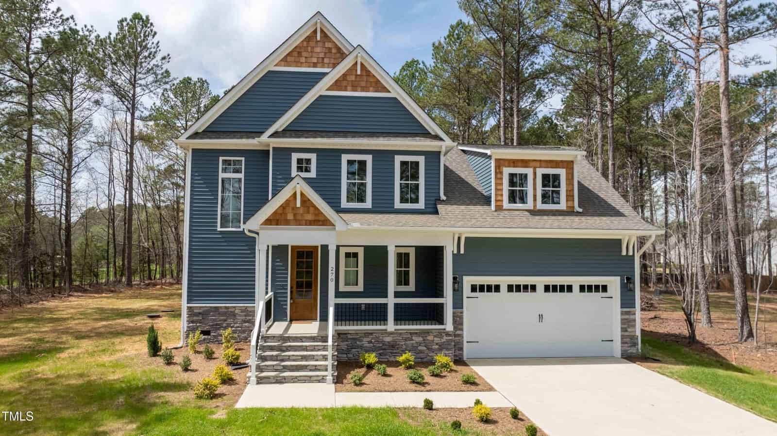 A two-story blue house with white trim, a covered front porch, and attached two-car garage is surrounded by trees. The front yard has small shrubs and a concrete driveway.