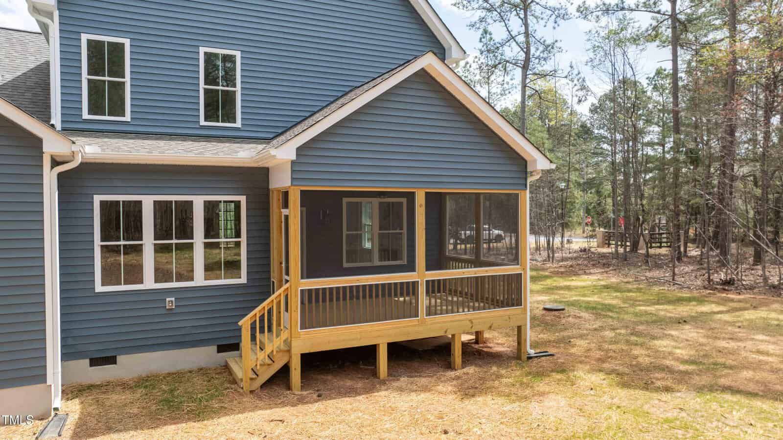 A blue house with white trim features a small screened porch with wooden steps and railing, set on a grassy yard bordered by trees.