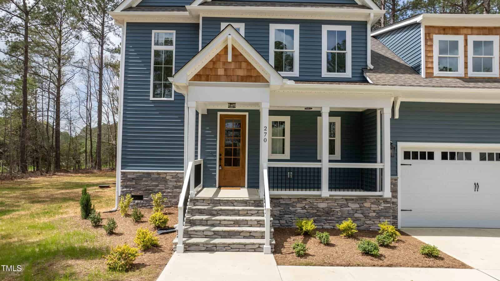 A two-story house with blue siding, stone accents, and a covered front porch. Steps lead to a wooden front door. There are shrubs and mulch along the walkway, and a white garage door is visible on the right.