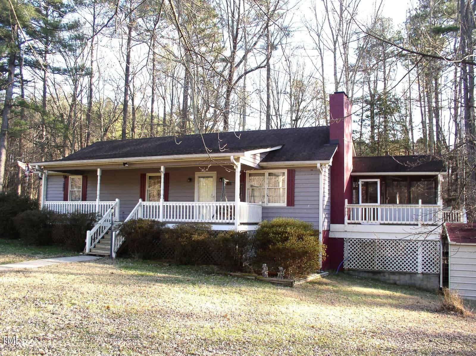 A one-story house on 105 Lake Road in Henderson features a covered front porch, painted light purple with white trim, surrounded by bare trees. The dark roof is complemented by a red brick chimney on the right side. A sloping yard leads up to the steps of the front entrance.
