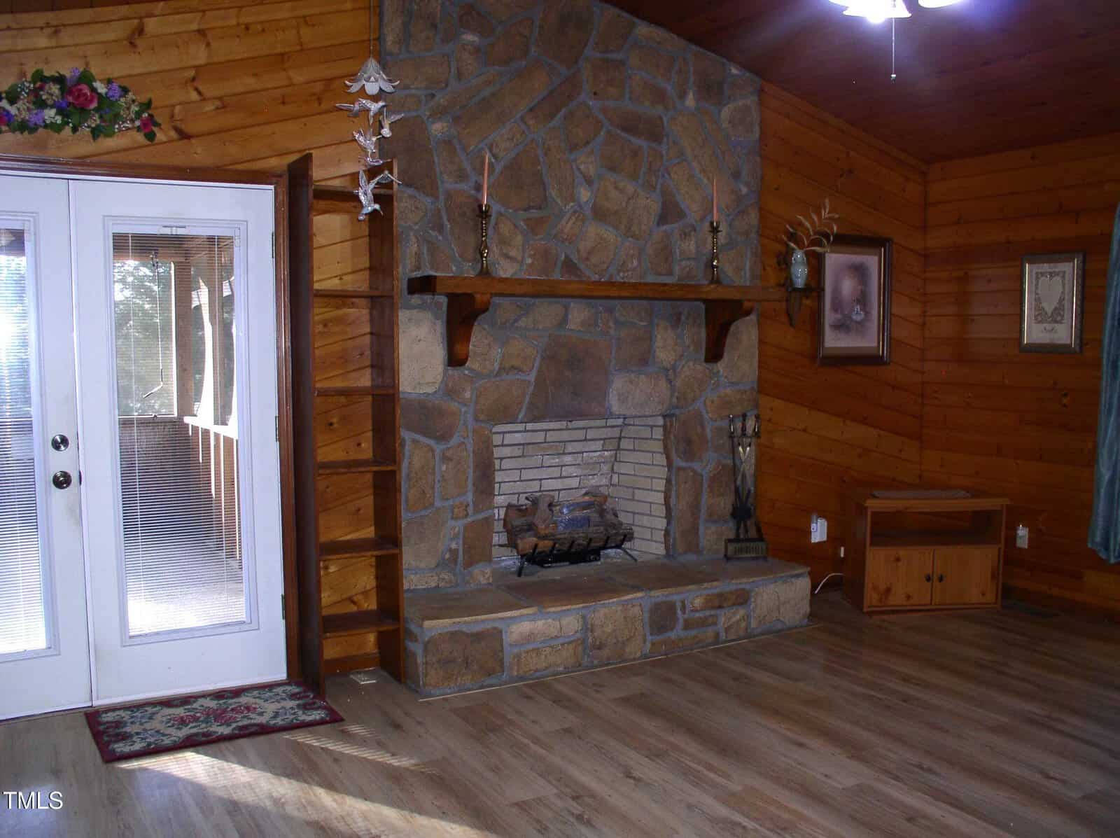 A room with wooden walls and floor features a large stone fireplace at its center. To the left are glass double doors with curtains, and decorations are on a shelf above the fireplace. A framed picture hangs to the right.