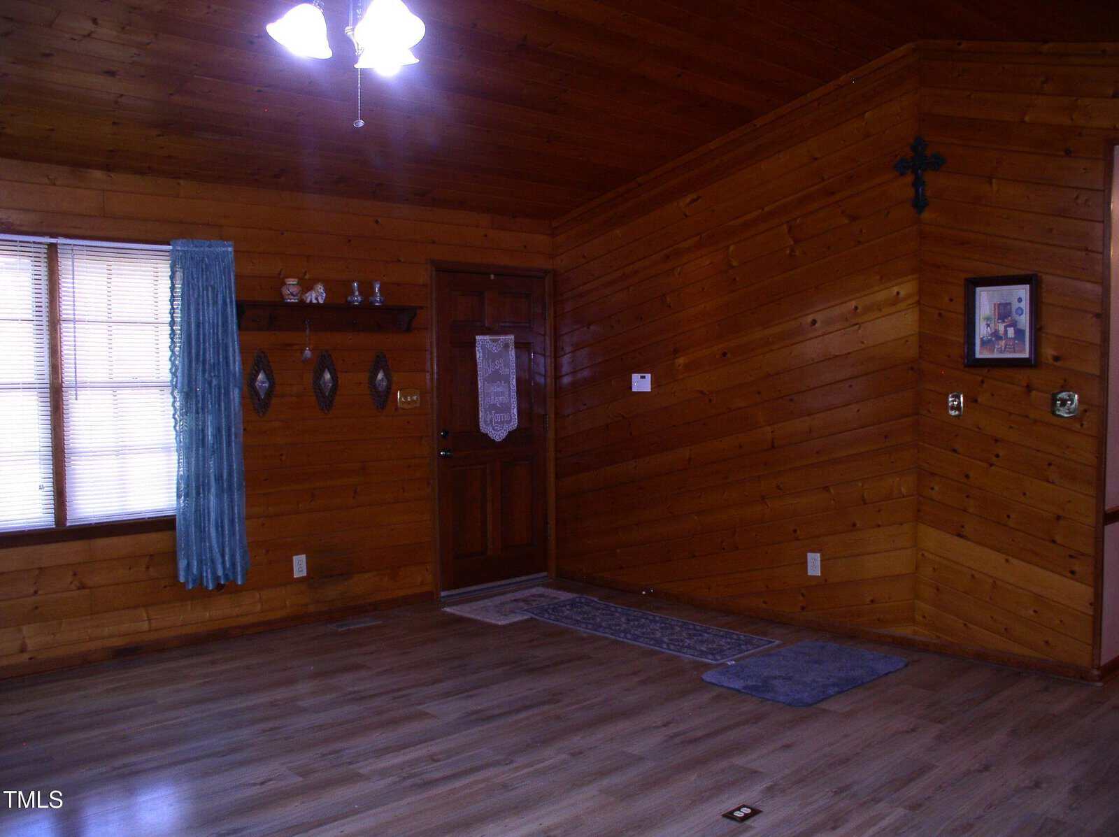 Wood-paneled room with hardwood floors, ceiling fan light, and window with blue curtains. Wall features small shelf with decor and a framed picture. Floor rugs near a wooden door with a hanging decoration.