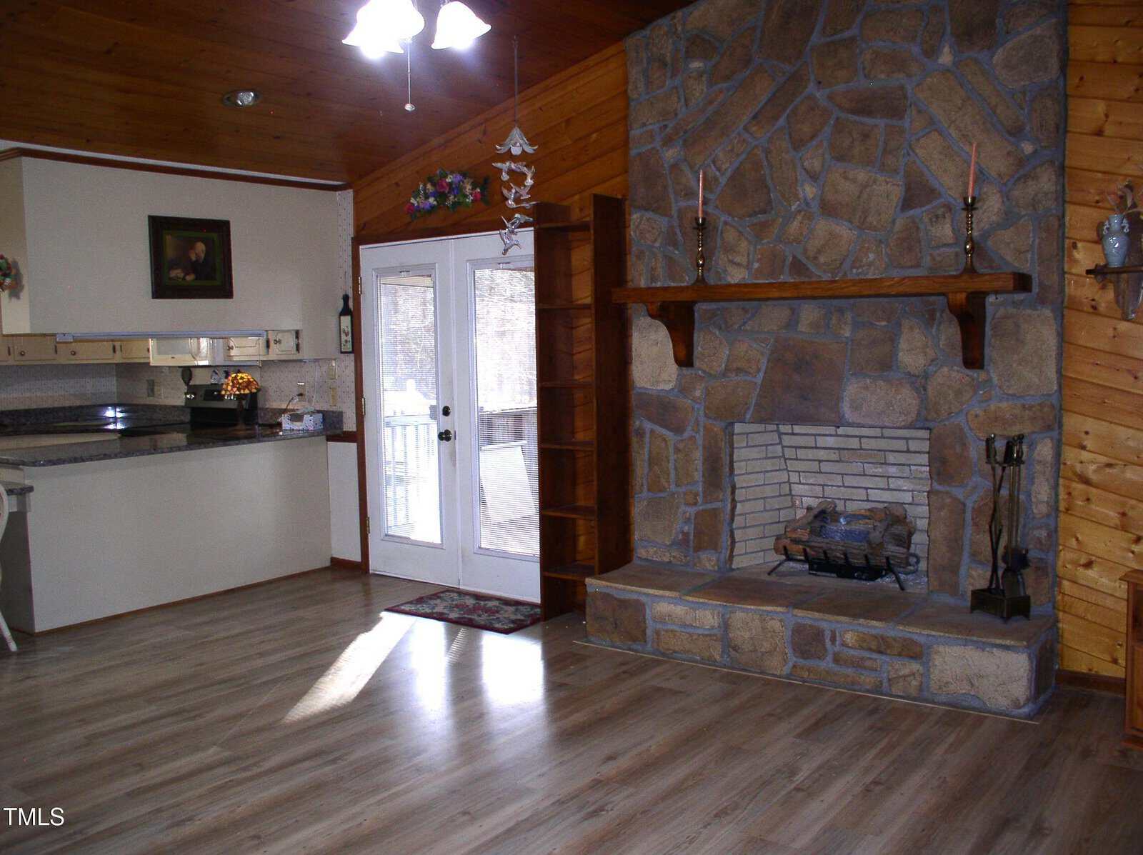 Interior of a room with a stone fireplace, wooden mantel, and built-in shelves. The room has wood-paneled walls and a ceiling fan. French doors lead to the outside. The floor is light wood, and a small kitchen area is visible on the left.