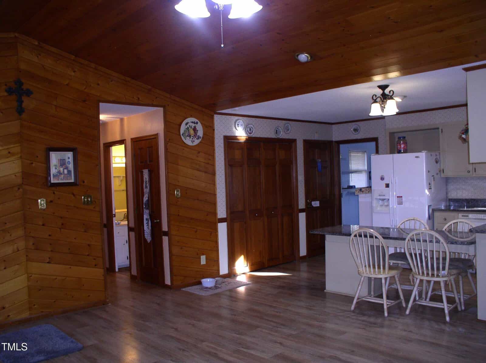 A kitchen and dining area with wooden paneling on the walls and ceiling. There is a white refrigerator, a dining table with four chairs, and wooden cabinets. A brightly lit hallway leads to an adjoining room. The flooring is light wood.