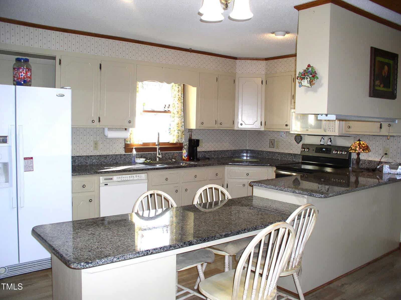 A kitchen with beige cabinets and granite countertops. There is an island with two wooden chairs and built-in appliances, including a refrigerator, stove, and dishwasher. A window with a floral curtain is above the sink, and a light fixture hangs from the ceiling.