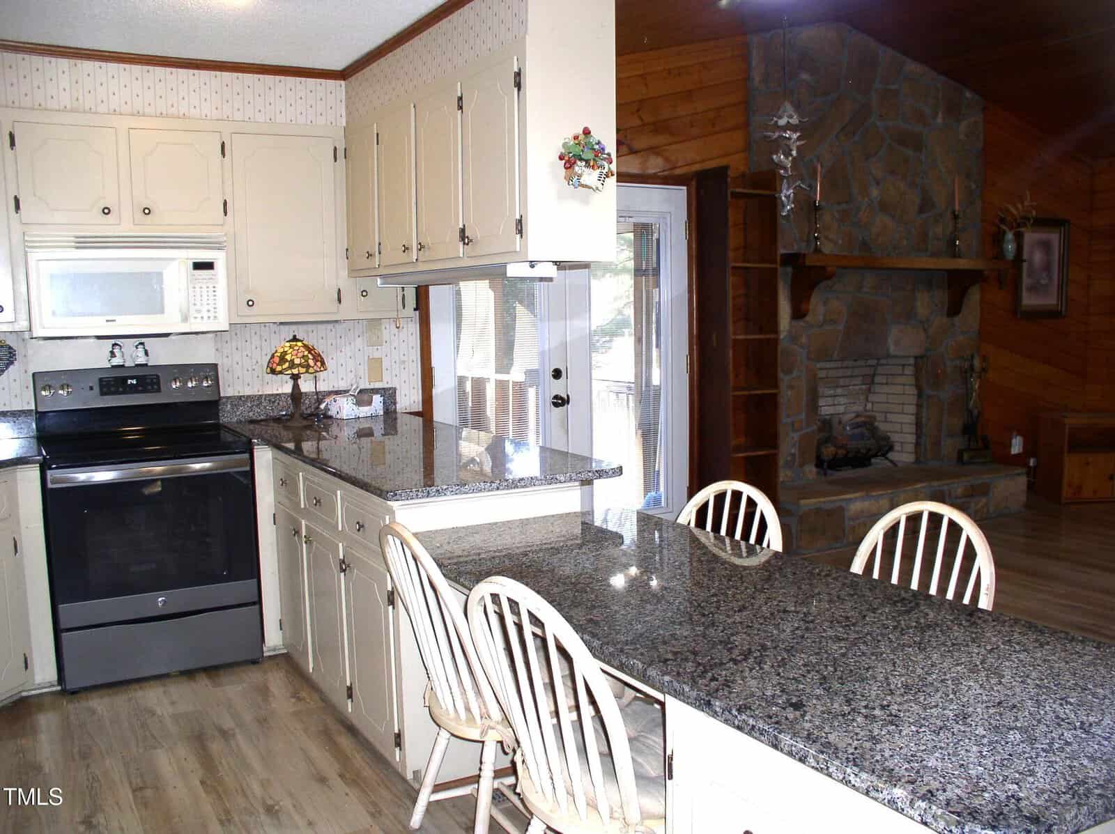 Kitchen and living area with wood paneling and stone fireplace. Kitchen features light-colored cabinets, granite countertops, and stainless steel appliances. A small dining area with three wooden chairs is integrated with the countertop.