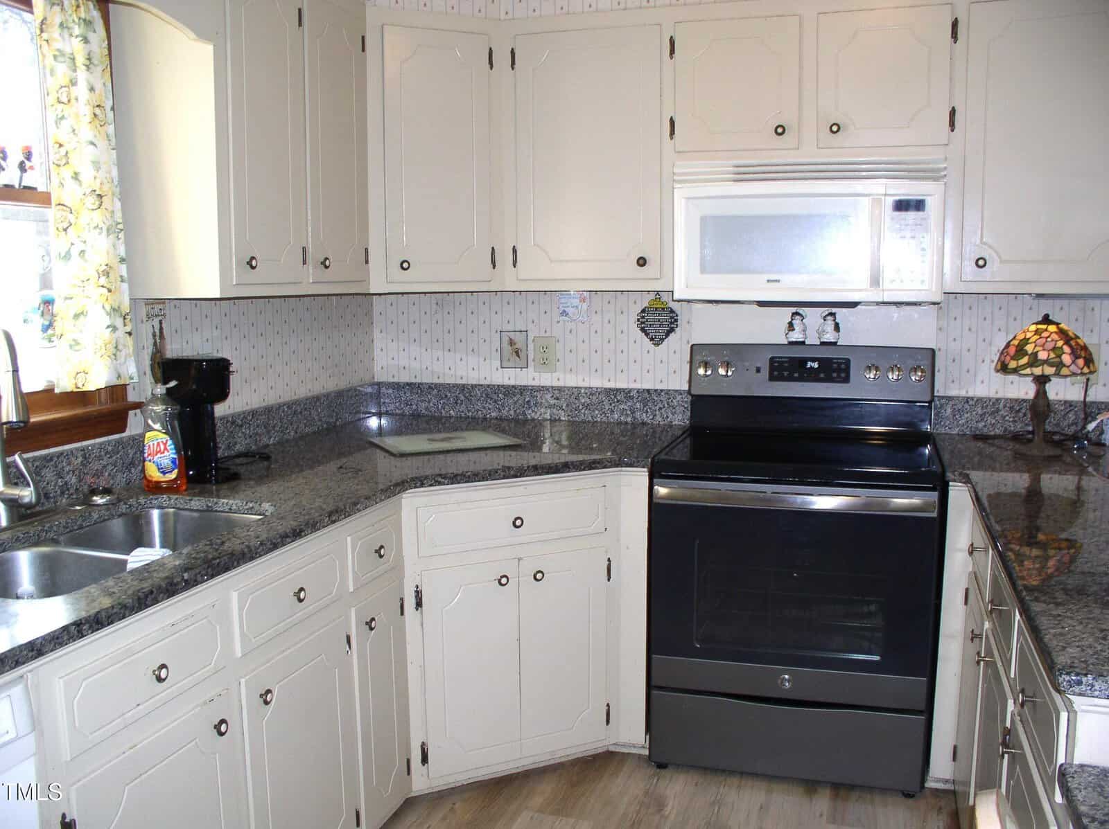 A kitchen with white cabinets, a granite countertop, and a black stove with an overhead microwave. A coffee maker is on the counter next to a sink. A small decorative lamp sits on the counter. The backsplash features a subtle patterned design.