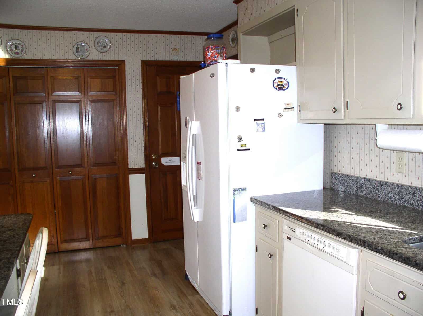 Kitchen interior featuring a white refrigerator, beige cabinets, and granite countertops. Wooden flooring and wall paneling complement the room. Decorative plates are displayed above a set of wooden double doors in the background.
