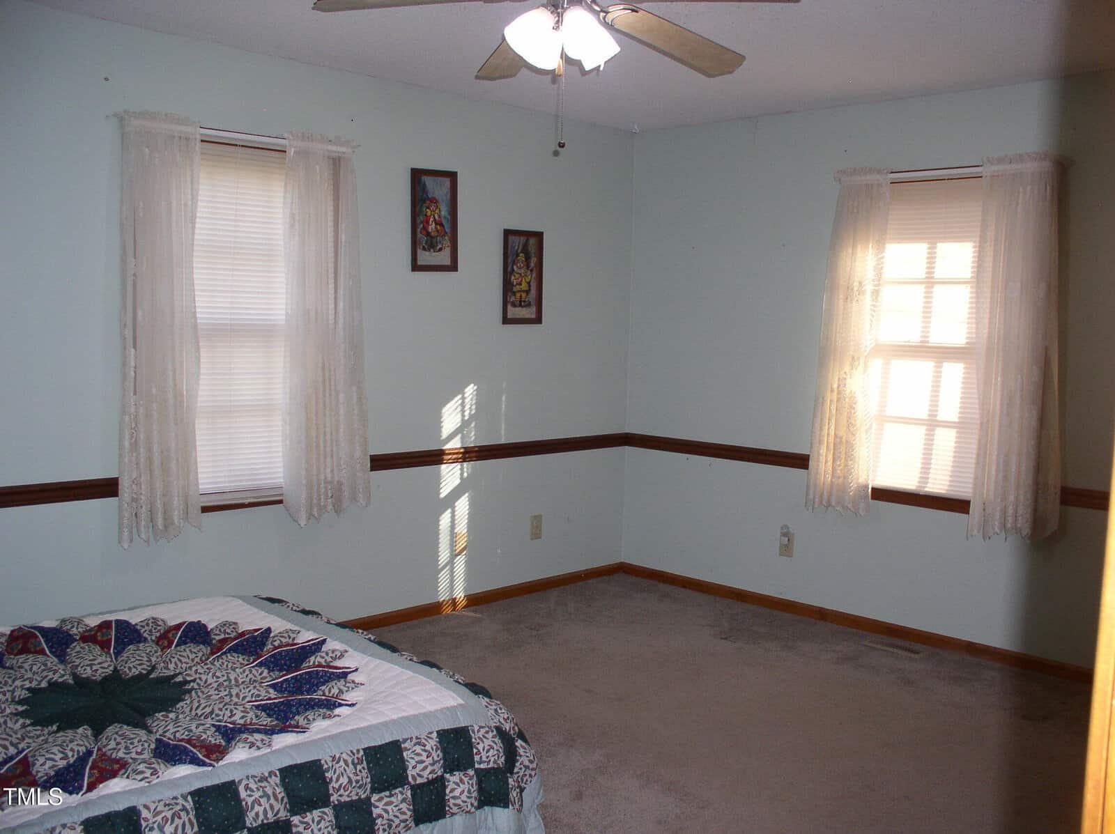A sparsely furnished bedroom with light blue walls, featuring two windows with lace curtains. There is a ceiling fan and a single bed with a patterned quilt. Two framed pictures hang between the windows. Light filters through the blinds.