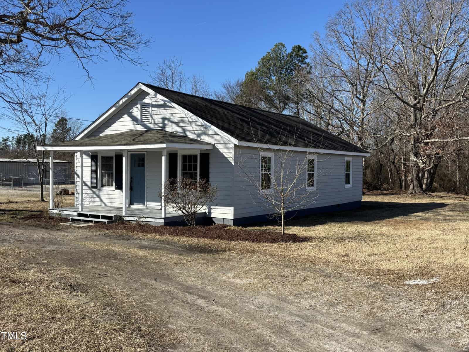 A small, white single-story house at 211 Satterwhite Point Road, Henderson, with a dark roof and a front porch, sits on a grassy plot. Leafless trees are in the background, and the sky is clear and blue. A dirt driveway leads to this charming home.