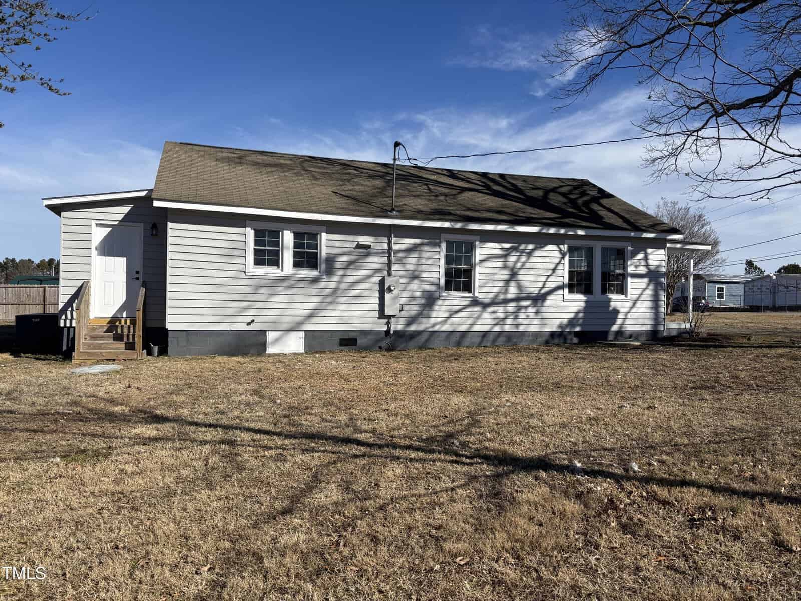 A single-story, light gray house with a gabled roof and white trim. It has several windows and a small set of steps leading to a white door. The house is surrounded by a dry grass yard, and the sky is clear and bright.