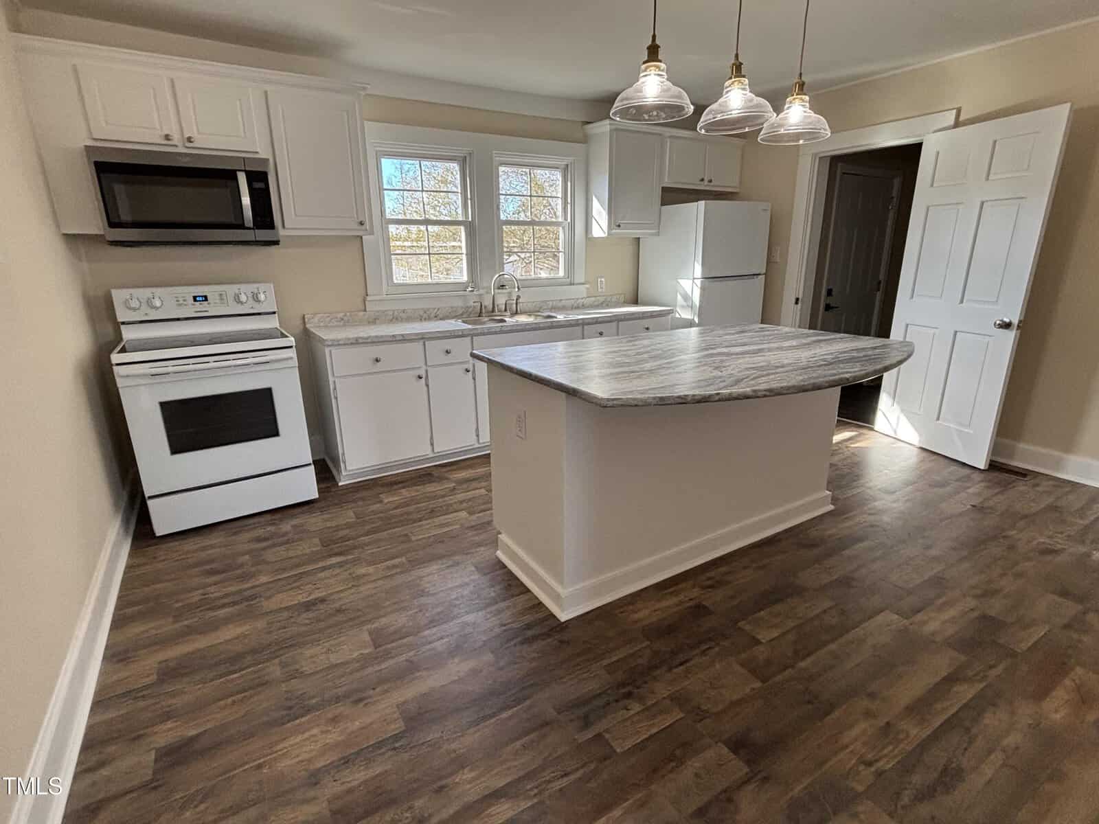 A kitchen with wooden flooring features a central island with a marble countertop. There is a white stove and oven, a microwave above it, a refrigerator, and cabinets. Three pendant lights hang above the island. Two windows are present above the sink.