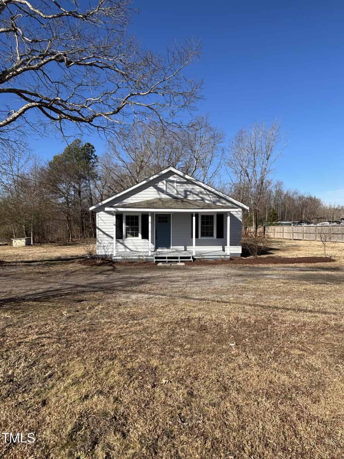 A small, white single-story house with a green front door and two windows. It sits on a grassy lot with sparse trees. A clear blue sky is overhead. A fence is visible on the right side of the property.