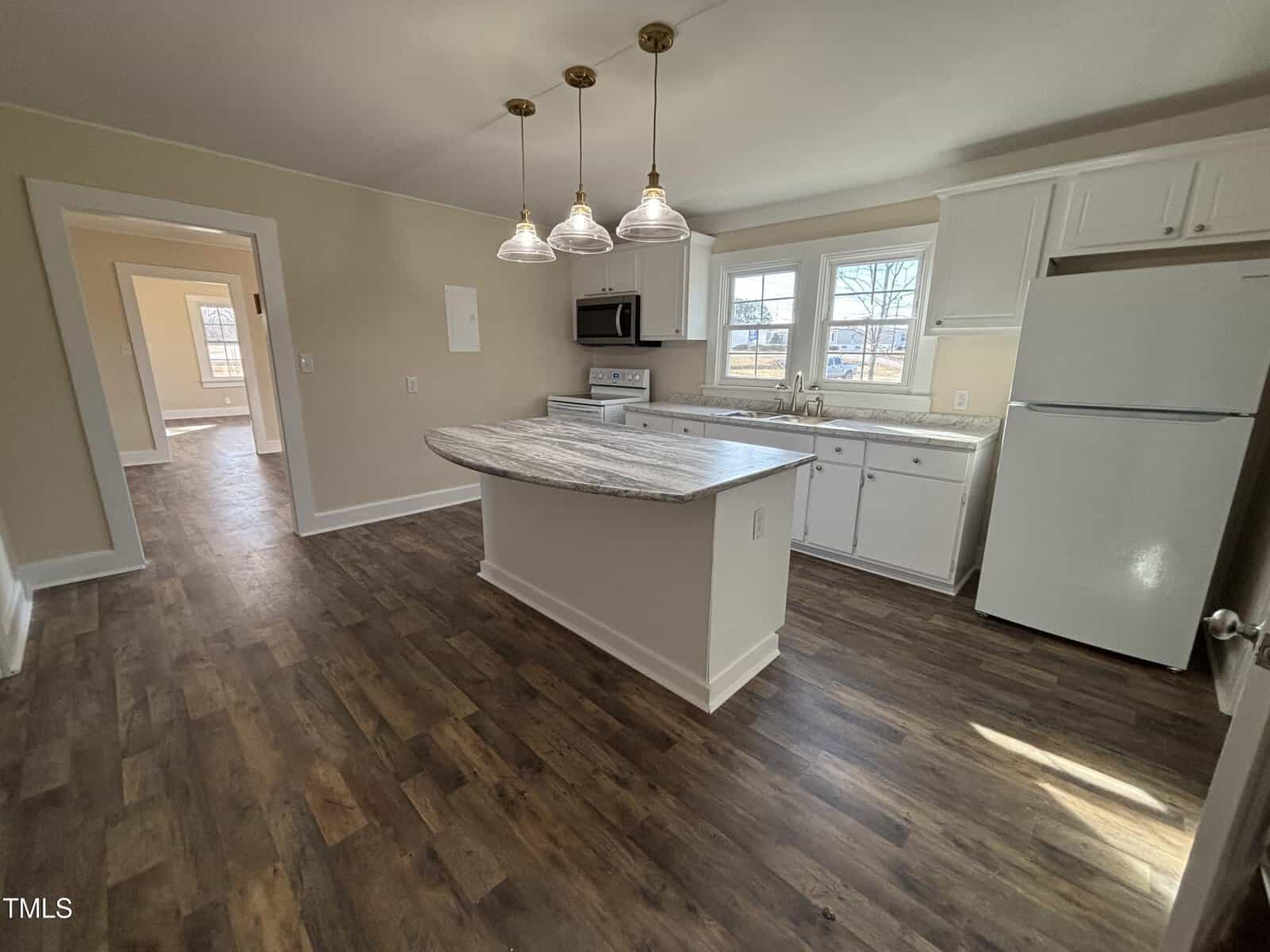Kitchen with wooden flooring and a central island with a speckled countertop. Pendant lights hang above the island. White cabinets, a refrigerator, and an oven are visible. Large windows offer natural lighting. Open doorway leads to another room.