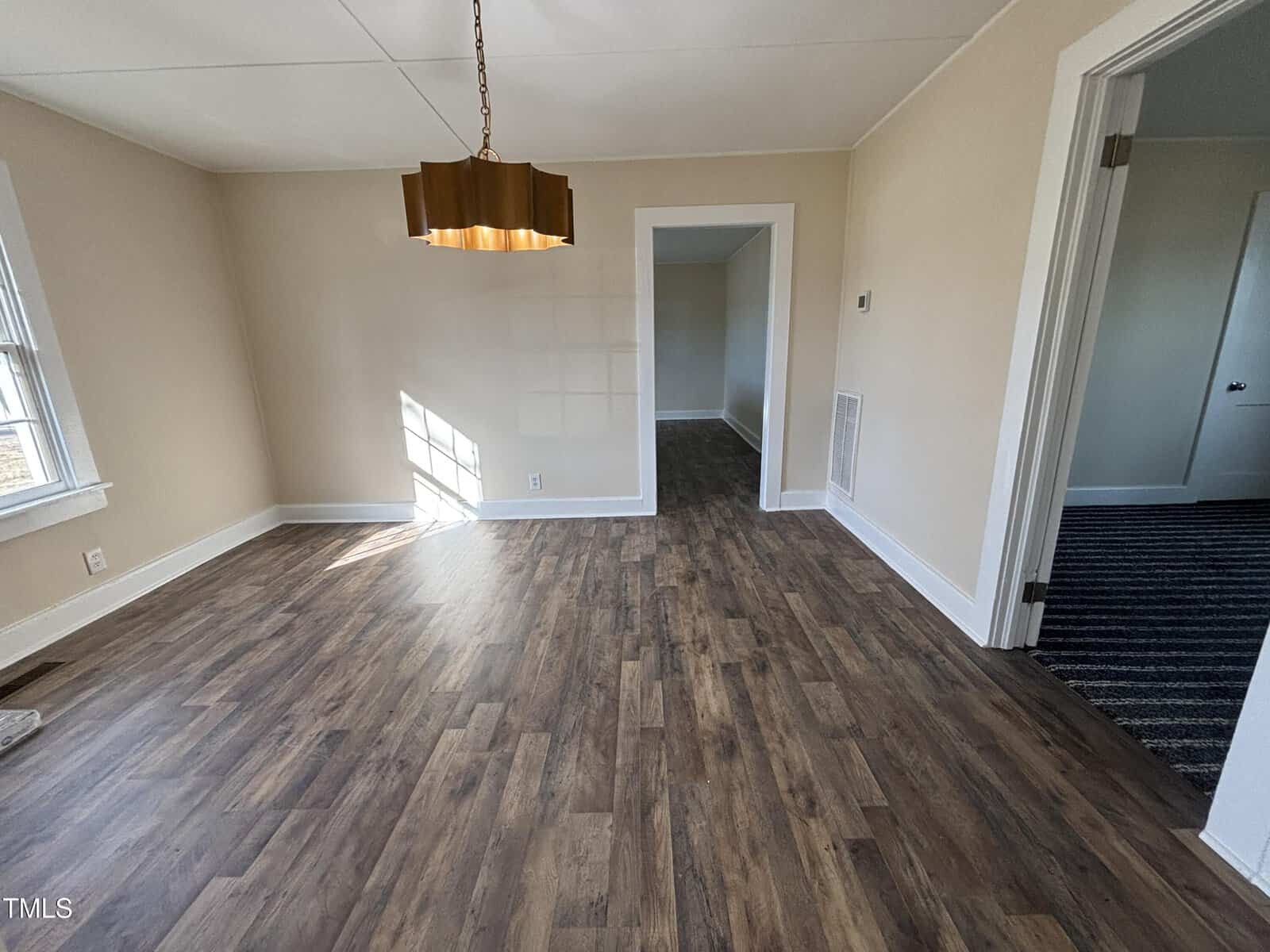 A bright, empty room with light brown wooden flooring and cream-colored walls. A large, geometric pendant light hangs from the ceiling. Sunlight streams through a window on the left, and adjacent rooms are visible through open doorways.