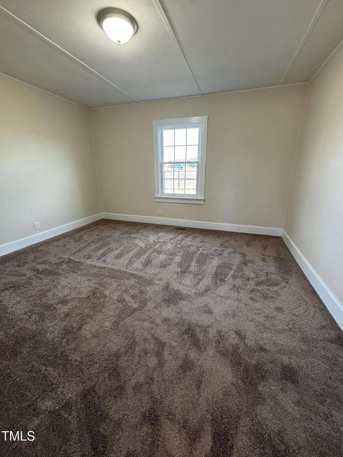 Empty room with beige walls and dark brown carpet. A small window centered on one wall lets in daylight. A ceiling light fixture is switched on. Skirting boards are painted white. Room appears freshly cleaned or vacuumed.