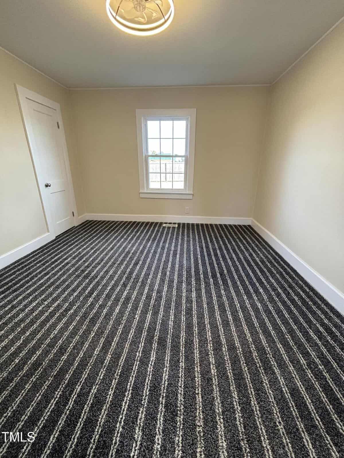 A small empty room with beige walls and a striped gray carpet. There is a single window in the center of the far wall, allowing natural light. A white door is on the left wall. The ceiling features a round, modern light fixture.