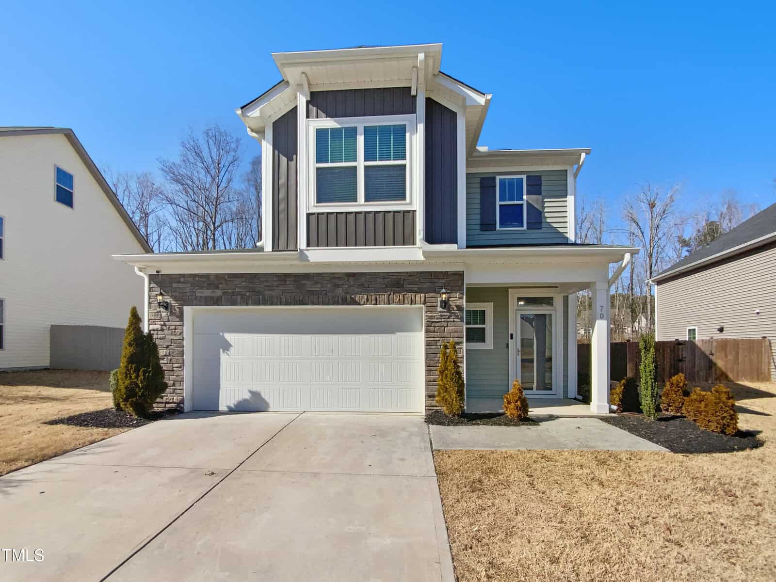 A two-story modern suburban house with gray siding, stone accents, white trim, and a double garage. The front yard has small shrubs and a wide concrete driveway, with neighboring houses visible on both sides.