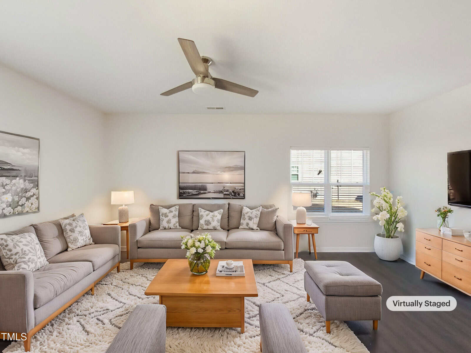 A bright living room with light gray walls, two beige sofas, a wooden coffee table, ottomans, a TV on a wooden console, wall art, lamps, and potted plants. A ceiling fan is mounted on the white ceiling.