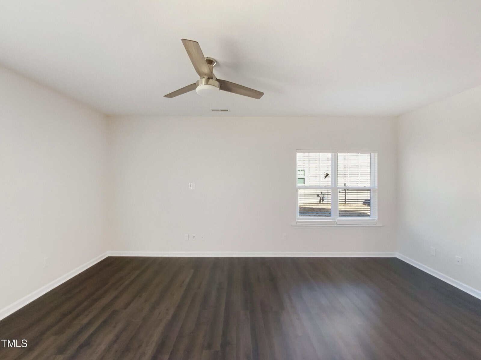 Empty room with white walls, dark wood flooring, a ceiling fan with light, and a window with white blinds letting in natural light.