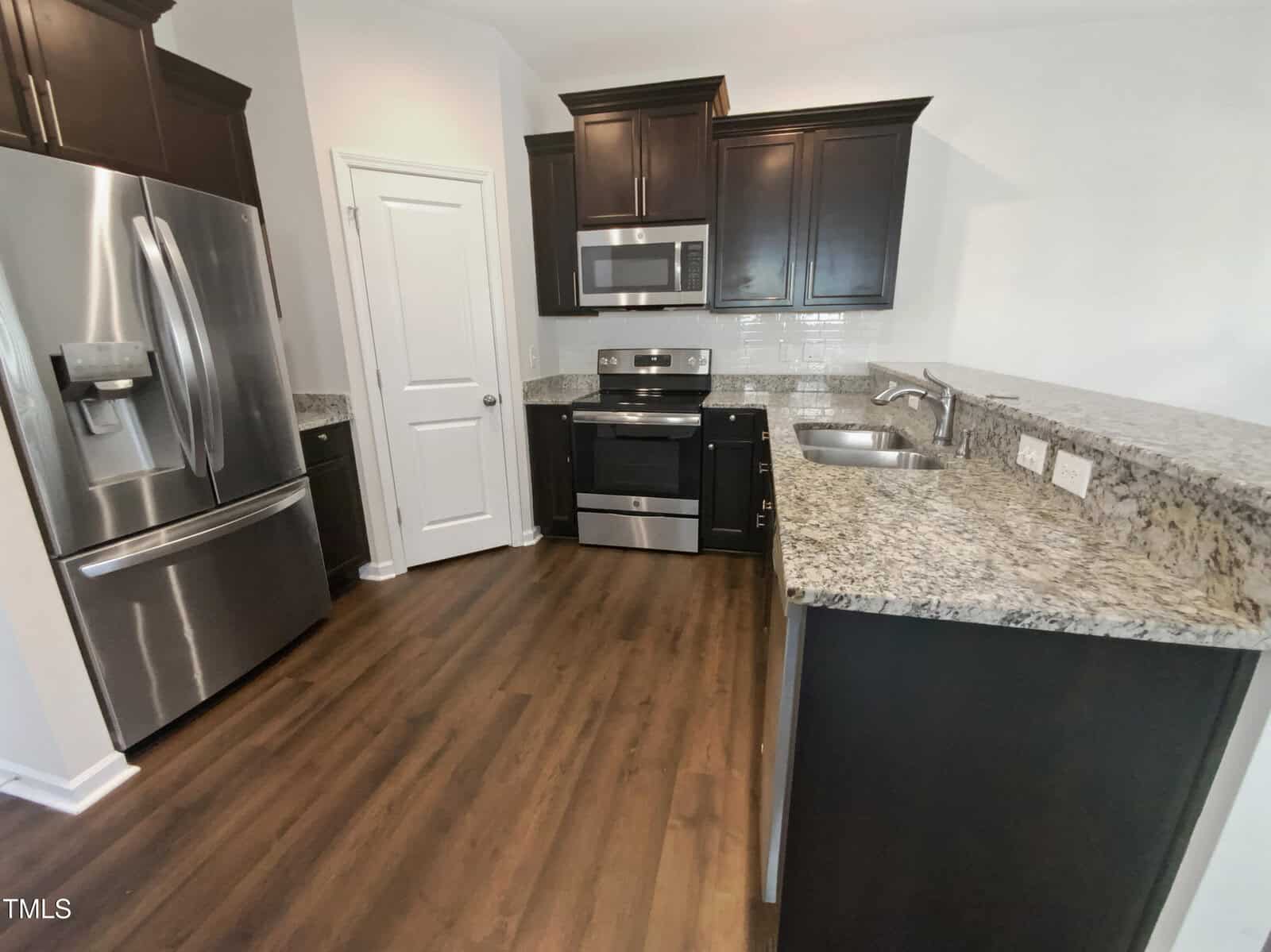 Modern kitchen with dark cabinets, stainless steel appliances, granite countertops, double sink, white tile backsplash, wood flooring, and a white door in the corner. Outlets are visible above the counter.