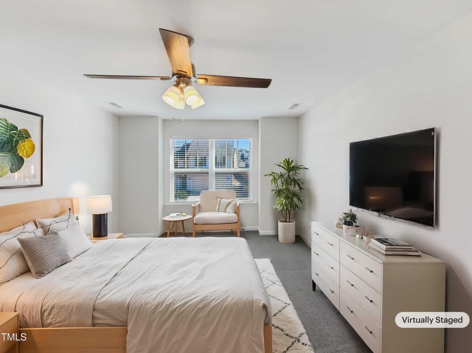 A bedroom with a bed, chair, dresser, TV, potted plant, and artwork. The room has neutral colors, a ceiling fan with lights, a window with blinds, and a rug. The bottom right corner has a “Virtually Staged” label.