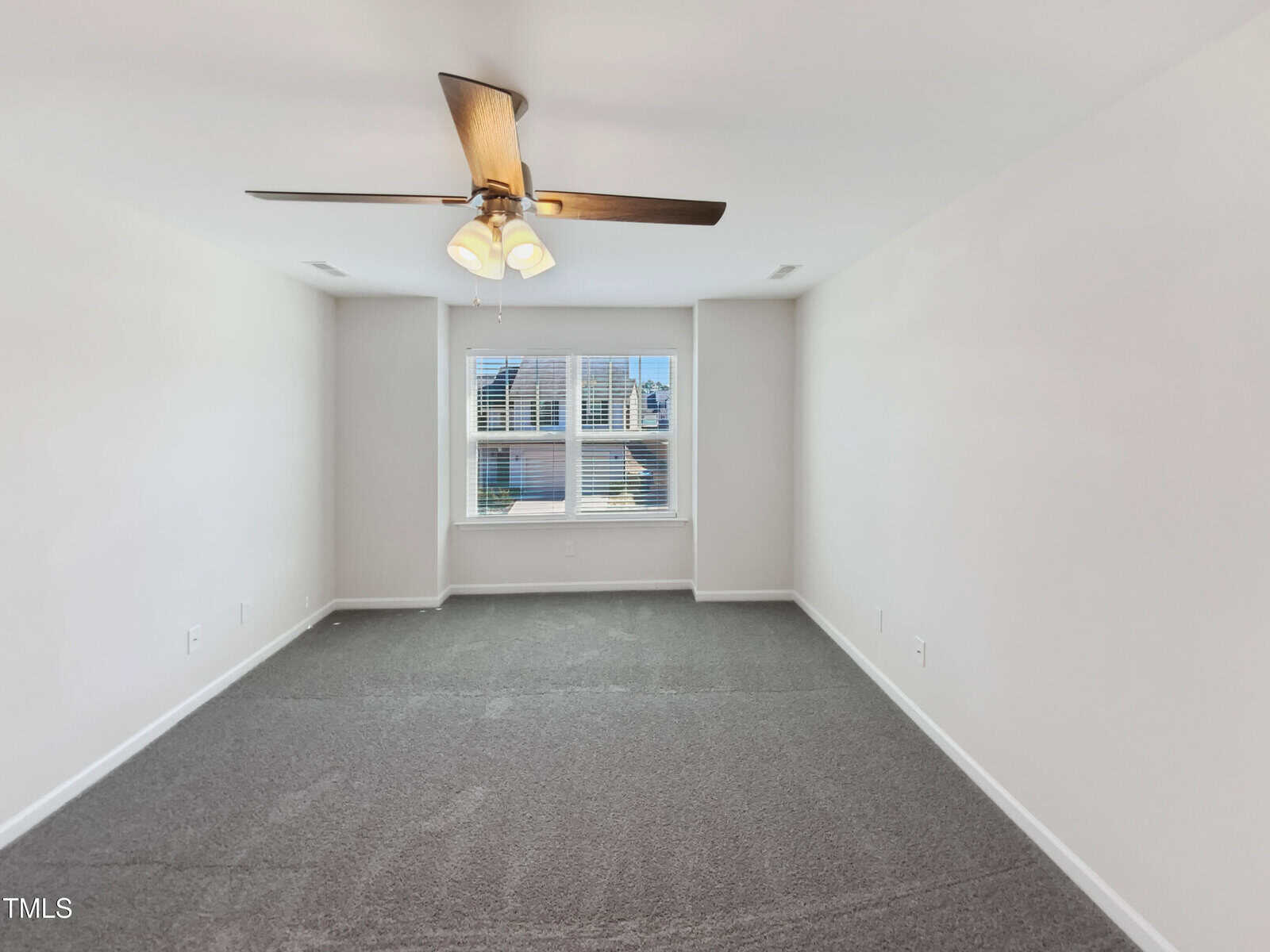 A vacant room with light gray carpet, white walls, a ceiling fan with lights, and a large window letting in natural light.
