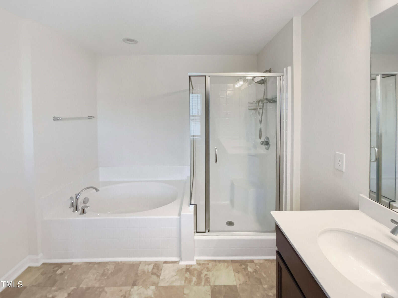 A bathroom features a white bathtub next to a glass-enclosed shower, with a towel bar on the wall. A vanity with a sink and countertop is visible in the foreground. The floor is tiled in a neutral color.