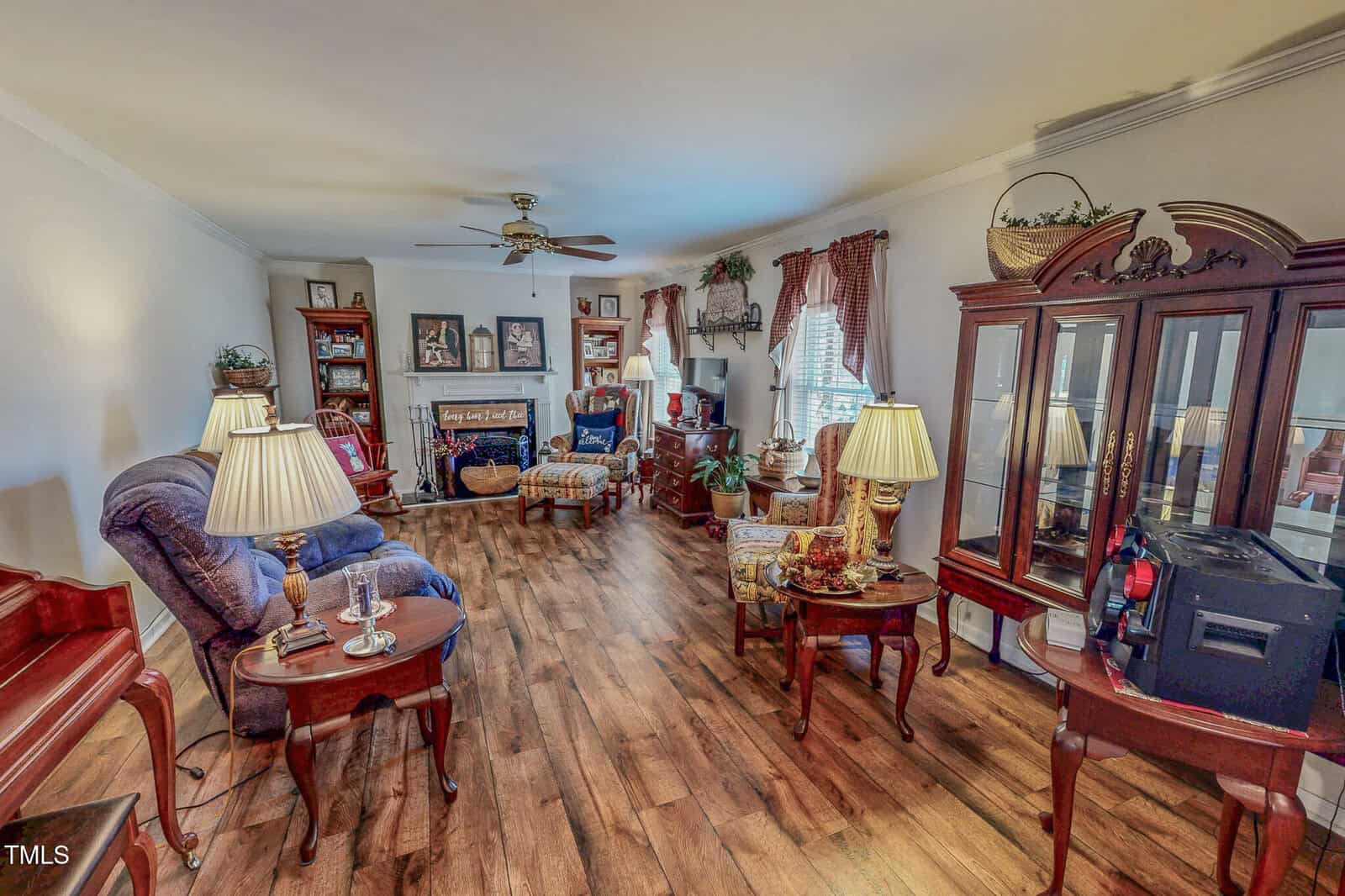 A cozy living room with wooden flooring and a variety of furniture, including chairs, a sofa, and side tables with lamps. A ceiling fan is above, and a large cabinet with glass doors is on the right. Wall art and plants decorate the space.