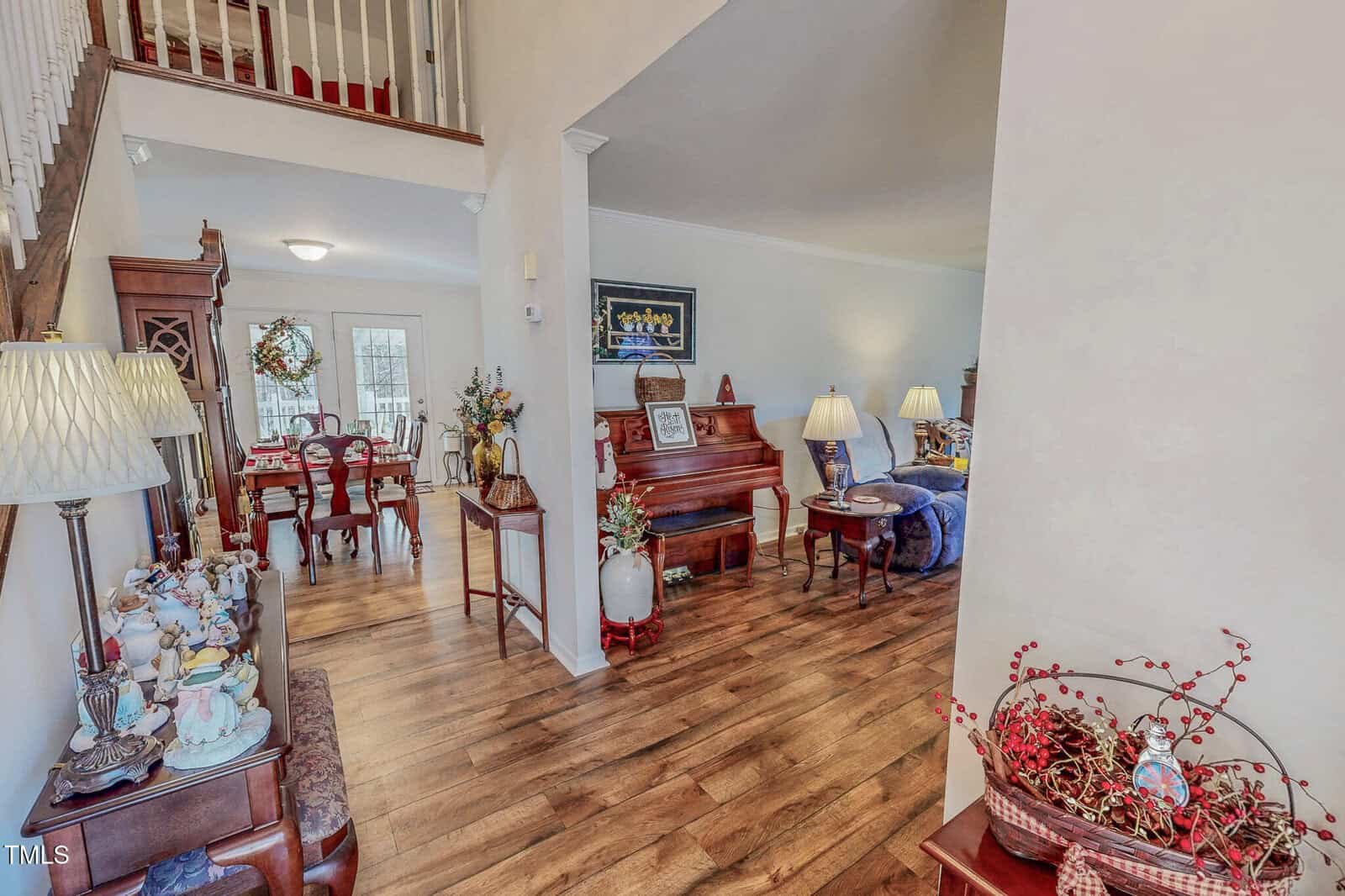 A cozy living room with wooden flooring features a piano, a couch, and a small table with a lamp. The room opens into a dining area with a neatly set table. A staircase leads to an upper floor. Decorative items are placed throughout the space.