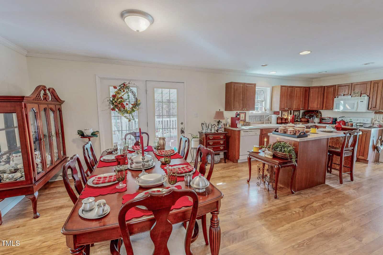 A dining room and kitchen area with wooden flooring. The dining table is set with red placemats, dishes, and silverware. A wreath hangs on glass doors. The kitchen features white appliances and wooden cabinets, with various items on the counters.