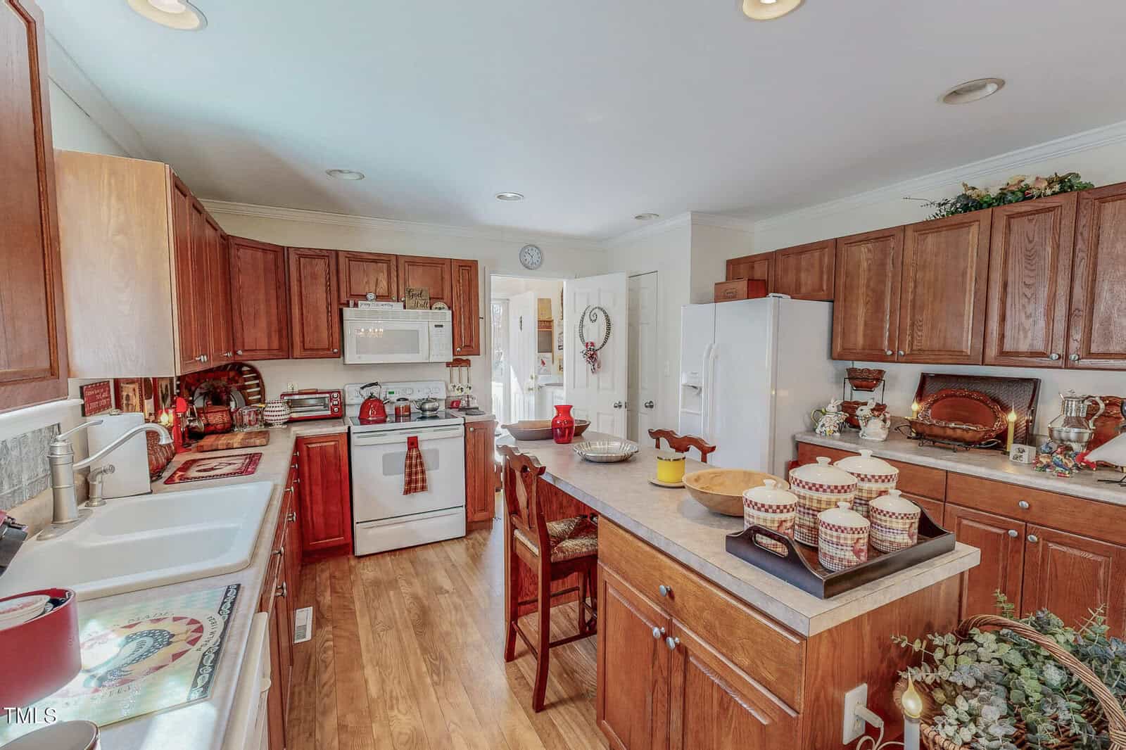 A kitchen with wooden cabinets and a central island. The countertop holds decorative bowls, mugs, and plants. Appliances include a white refrigerator, stove, and microwave. The room features hardwood flooring and ample natural light.
