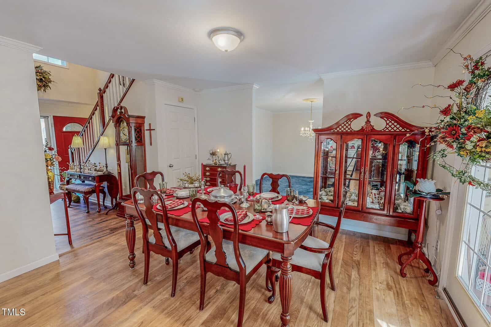 A dining room with a wooden table set for six, featuring red cushioned chairs and tableware. There's a china cabinet against the wall and decorative floral arrangements. The room has hardwood floors and an adjoining hallway with stairs.