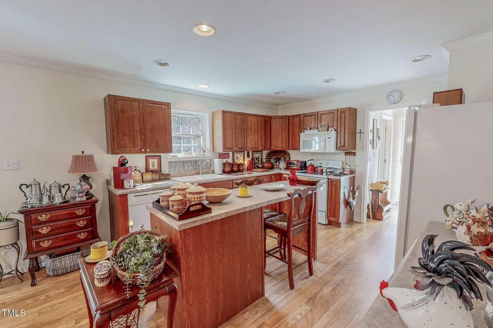 A kitchen with wooden cabinets and a central island. Two chairs are at the island, which holds bowls and decor. Appliances include a stove, microwave, and refrigerator. A small lamp and decor items are placed on a side table. A plant sits on a chair.