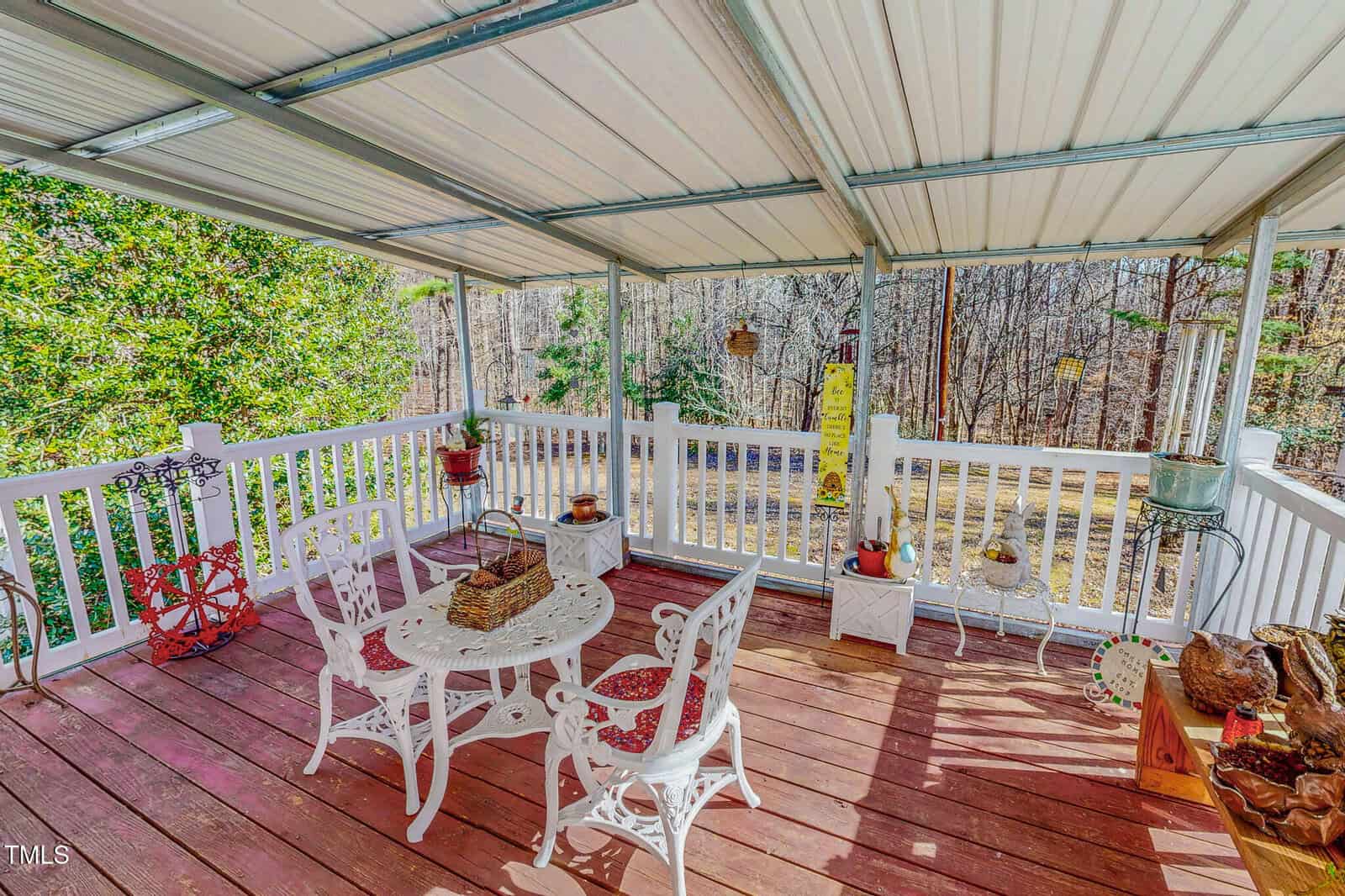 A covered patio with a wooden floor features a white metal table and two chairs. The table has a basket centerpiece. Potted plants and decor surround the area. A bird feeder is visible in the background amid trees and foliage.
