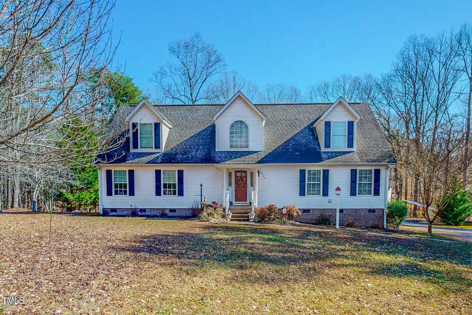 Located at 3134 Magnolia Lane, this charming two-story house boasts a grey gabled roof, white siding, and blue shutters. A large window elegantly graces the entrance. In the backdrop, leafless trees stand tall, while dried leaves and grass blanket the ground with an autumnal touch.